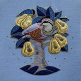 A square textile artwork or embroidered cushion featuring a partridge perched on a tree with blue leaves and gold seed pods on a light blue fabric background.