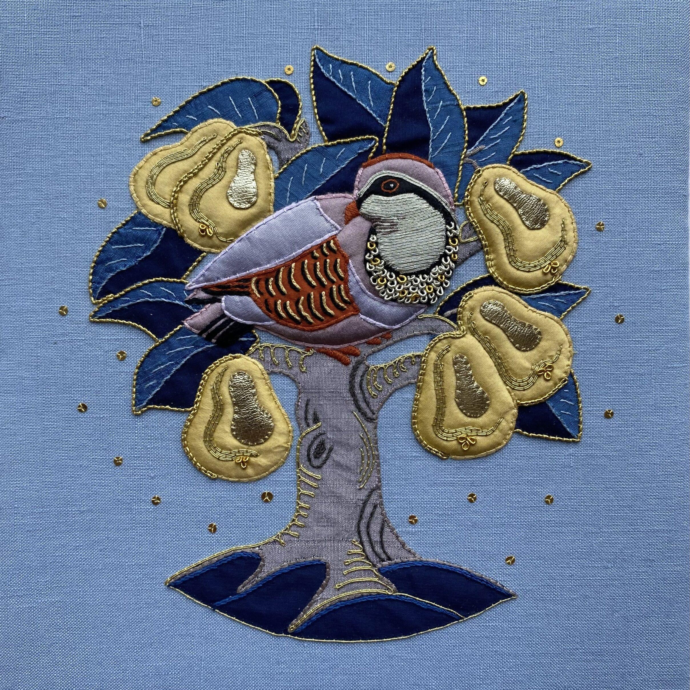 A square textile artwork or embroidered cushion featuring a partridge perched on a tree with blue leaves and gold seed pods on a light blue fabric background.