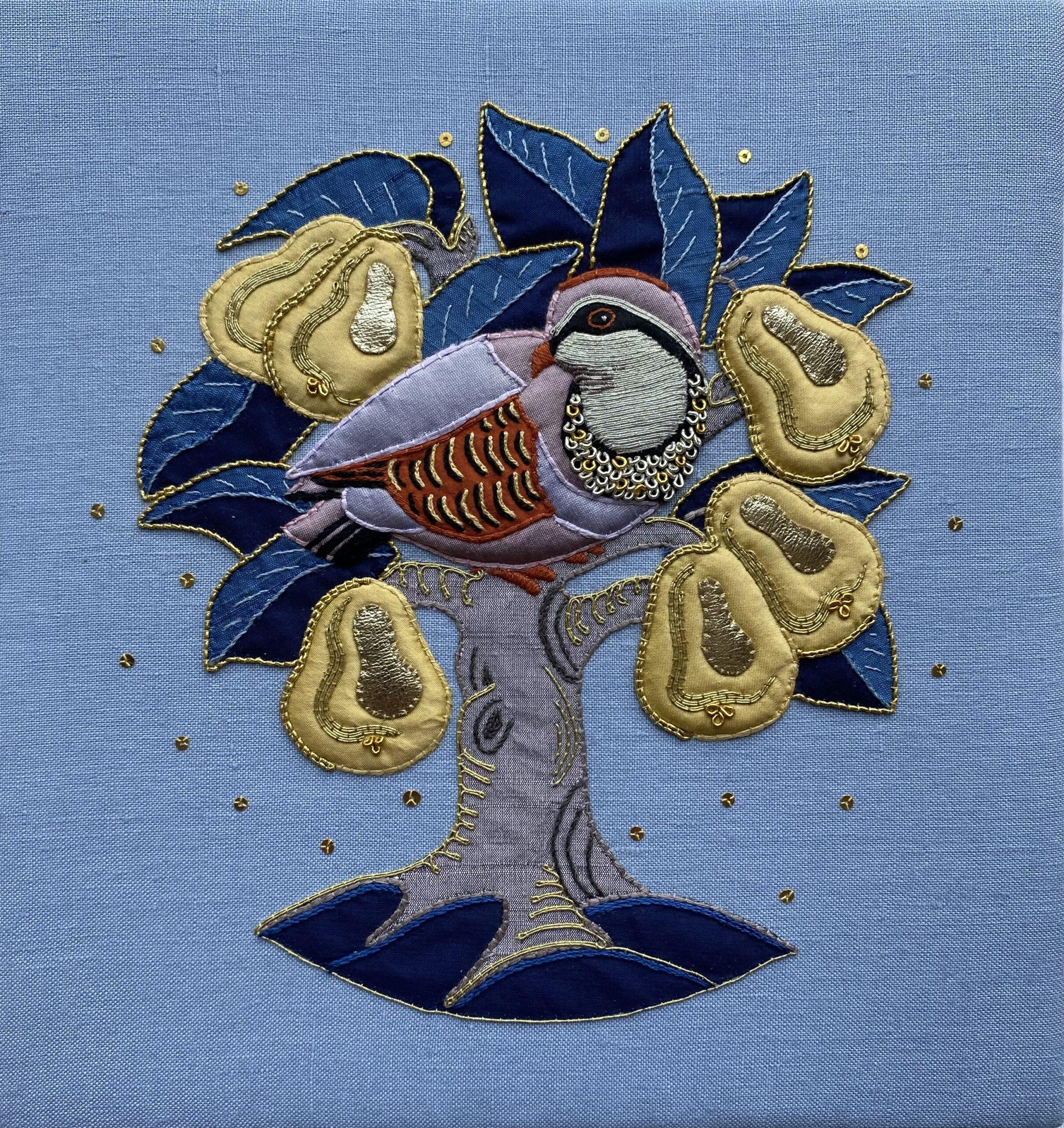 A square textile artwork or embroidered cushion featuring a partridge perched on a tree with blue leaves and gold seed pods on a light blue fabric background.